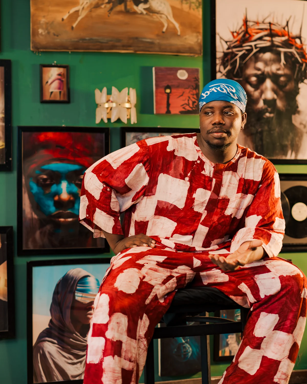 Deji Alaka seated in a studio filled with paintings, wearing a red and white adire two-piece